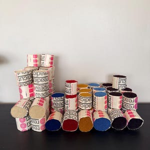 May include: A collection of cylindrical Readicut rug yarn bundles in various colors including red, blue, yellow, and dark purple. Each bundle is wrapped in a white paper band featuring the black text Readicut and pink circular patterns.