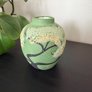 May include: A round ceramic vase with a matte sage green finish. The exterior features a black painted tree silhouette with textured, raised pale yellow and light green crackle glaze detailing that mimics foliage across the upper section.