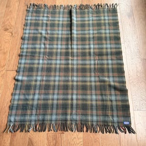 May include: A rectangular throw blanket featuring a plaid pattern in shades of dark green, brown, light blue, and thin yellow lines. The blanket has a fringed edge along the top and bottom sides and a small rectangular label in the corner.