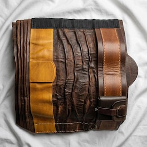 May include: A handcrafted leather tool roll in shades of brown and tan, featuring a black fabric top. The roll includes a small, attached pouch and a leather strap with a buckle. The leather has a textured, worn appearance.