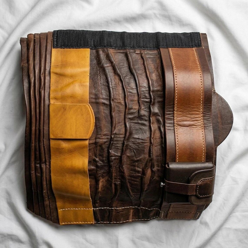 May include: A handcrafted leather tool roll in shades of brown and tan, featuring a black fabric top. The roll includes a small, attached pouch and a leather strap with a buckle. The leather has a textured, worn appearance.