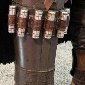 May include: Close-up of a brown leather leg armor with silver cylindrical canisters secured by brown leather straps. The armor is part of a costume, with a black fabric visible in the background.