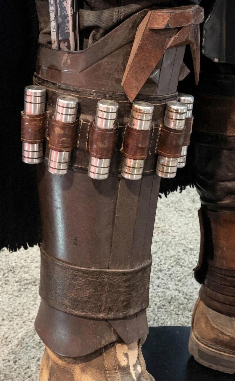 May include: Close-up of a brown leather leg armor with silver cylindrical canisters secured by brown leather straps. The armor is part of a costume, with a black fabric visible in the background.
