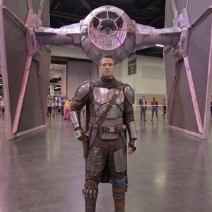 May include: A person in a Mandalorian costume stands in front of a large TIE fighter model. The costume features a silver chest plate, brown and gray armor, and a utility belt. The TIE fighter is gray and black.