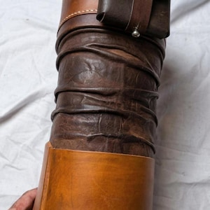 May include: A handcrafted leather item, possibly a container or sheath, in shades of brown. The design features a cylindrical body with a textured, wrinkled appearance, accented by lighter brown bands and a small, attached pouch.
