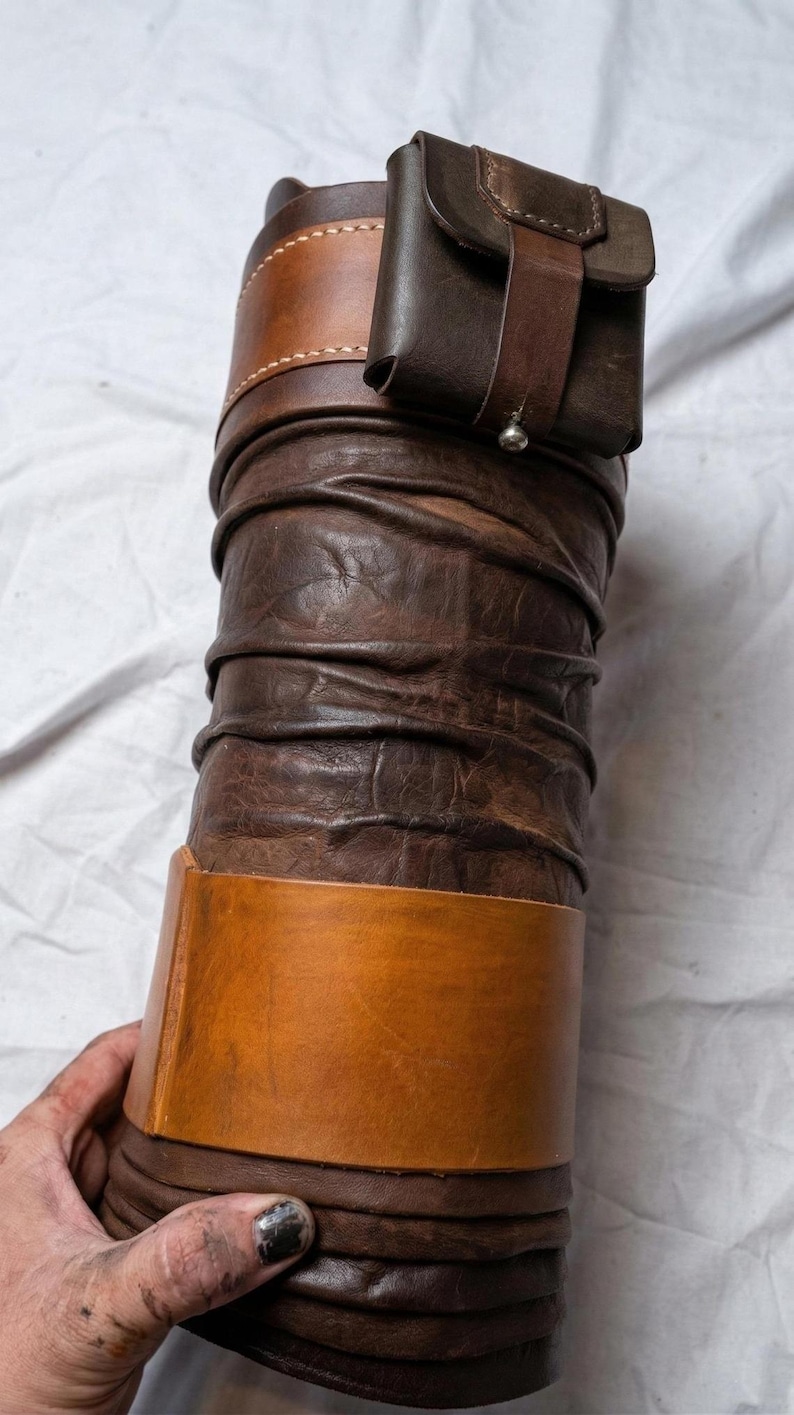 May include: A handcrafted leather item, possibly a container or sheath, in shades of brown. The design features a cylindrical body with a textured, wrinkled appearance, accented by lighter brown bands and a small, attached pouch.