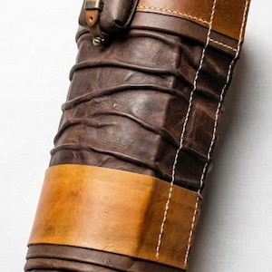 May include: A brown leather arm guard with a small pouch attached. The arm guard features a layered design with stitched accents and a contrasting tan leather band. The pouch has a buckle closure. The overall design is rugged and functional.