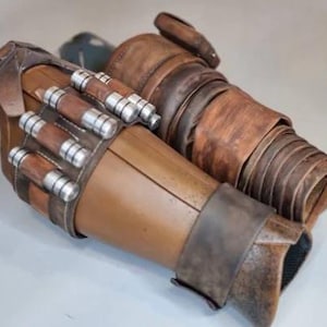 May include: A pair of brown and tan arm guards with silver and brown cylindrical details. The arm guards have a weathered appearance, with visible stitching and layered textures. The design suggests a futuristic or fantasy theme.