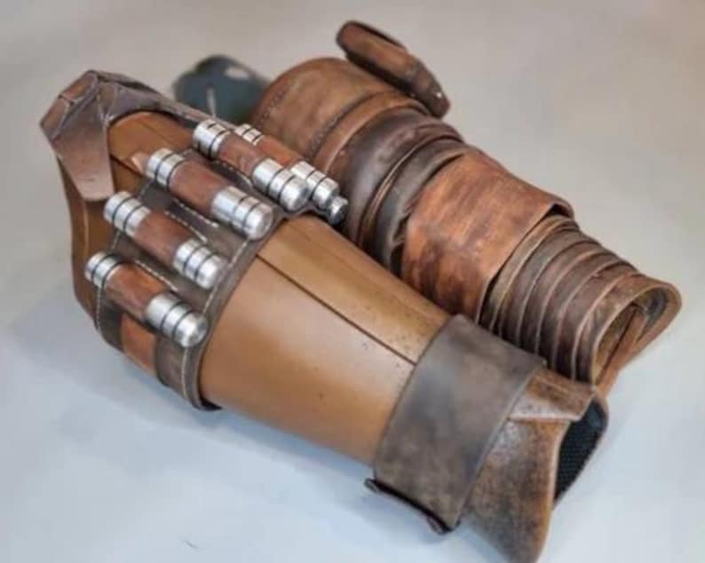 May include: A pair of brown and tan arm guards with silver and brown cylindrical details. The arm guards have a weathered appearance, with visible stitching and layered textures. The design suggests a futuristic or fantasy theme.