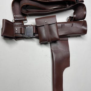 May include: A brown leather gun belt with multiple cartridge holders, a pouch, and a holster. The belt features a silver-colored buckle and is designed to be worn around the waist. The leather appears to be stitched for durability.