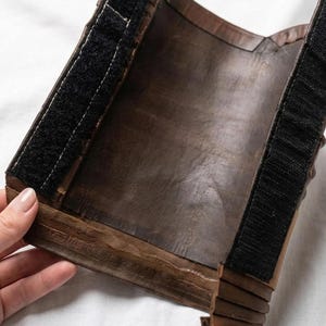 May include: A brown leather wrist cuff with a black velcro strap. The cuff is made of multiple layers of leather and has a textured appearance. The cuff is designed to wrap around the wrist.