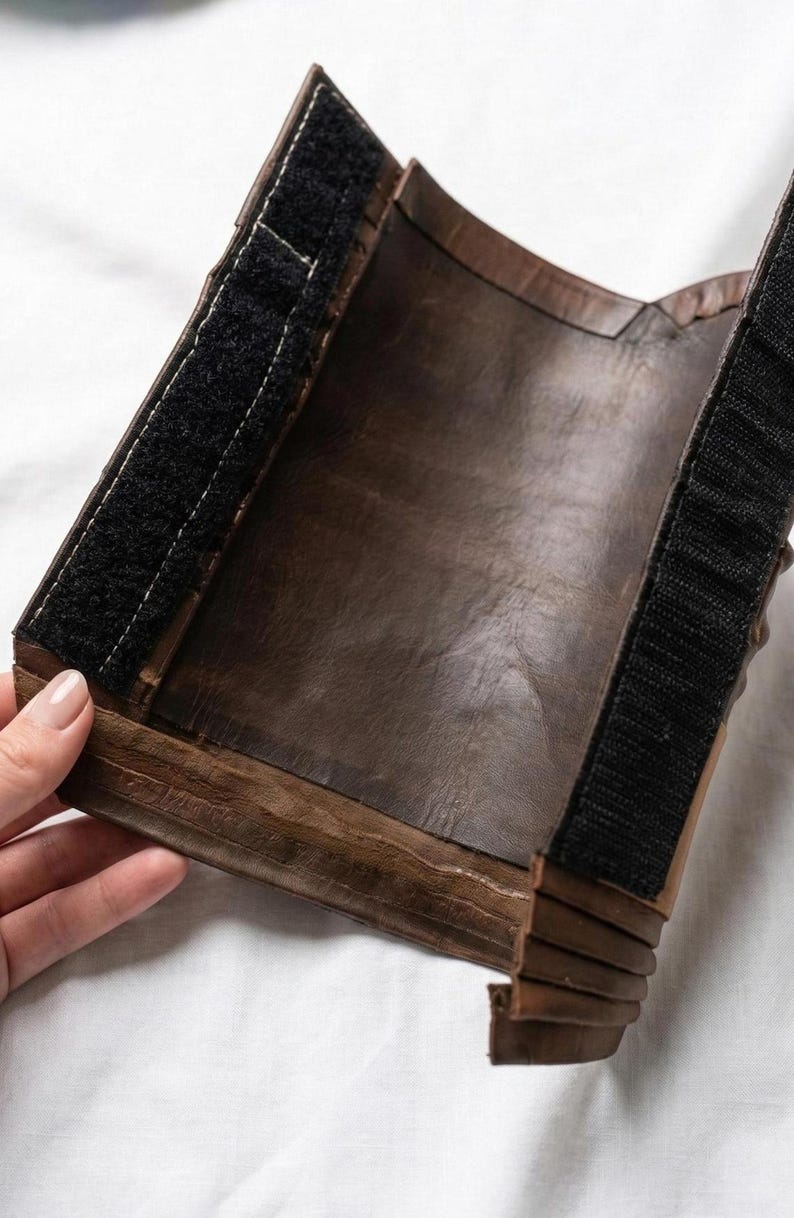 May include: A brown leather wrist cuff with a black velcro strap. The cuff is made of multiple layers of leather and has a textured appearance. The cuff is designed to wrap around the wrist.