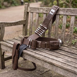 May include: A brown leather utility belt with multiple pouches, cartridge holders, and a silver-colored canister. The belt is resting on a wooden bench outdoors. The belt has a wide shoulder strap and a rectangular metal plate.