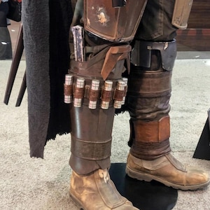May include: Close-up of a costume's lower half, featuring weathered brown boots and leg armor. The armor includes a holster with a silver object and a series of cylindrical containers. The costume is on display, with a dark cape visible on the left.