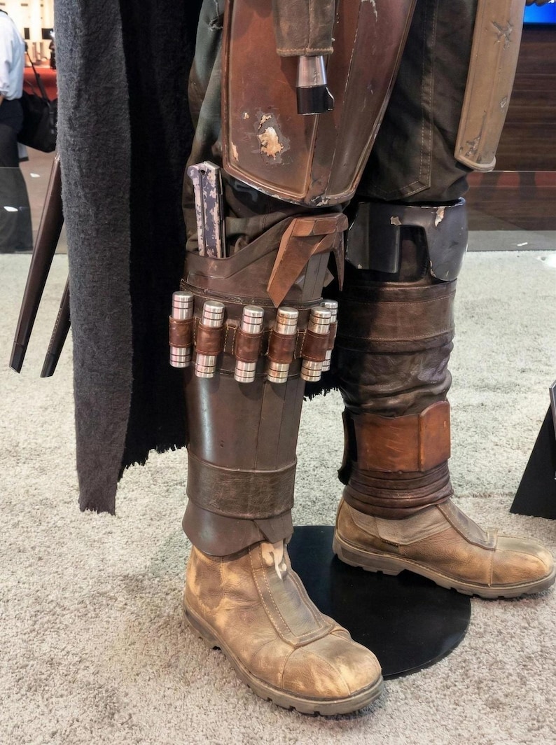 May include: Close-up of a costume's lower half, featuring weathered brown boots and leg armor. The armor includes a holster with a silver object and a series of cylindrical containers. The costume is on display, with a dark cape visible on the left.