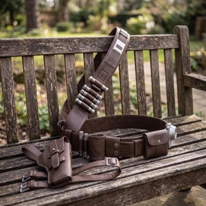 May include: A brown leather belt and holster set with silver accents. The set includes a wide belt with cartridge holders, a shoulder strap, a holster, and a small pouch. The items are displayed on a wooden bench.