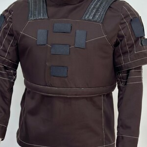 May include: Brown uniform with black accents, featuring a high collar and reinforced shoulders. The outfit includes a jacket with rectangular patches and matching pants. White stitching outlines the seams and details.