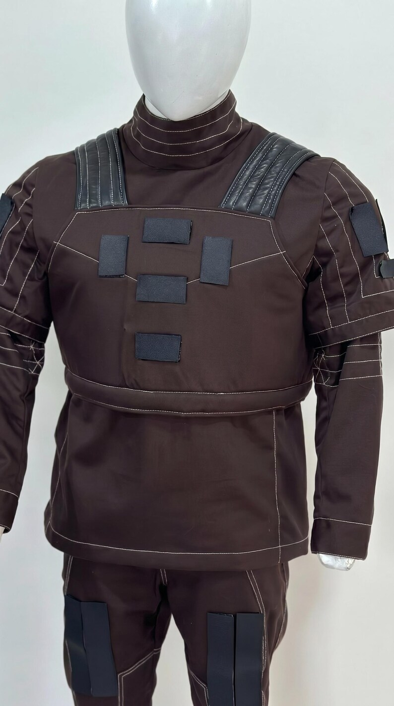 May include: Brown uniform with black accents, featuring a high collar and reinforced shoulders. The outfit includes a jacket with rectangular patches and matching pants. White stitching outlines the seams and details.