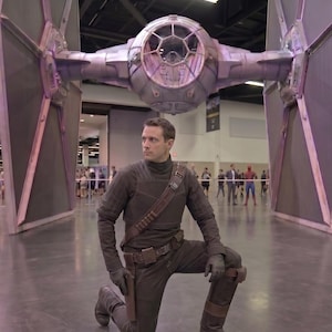 May include: A person kneels in front of a large, detailed model of a TIE fighter. The person is wearing a dark brown costume with a holster and gloves. The TIE fighter is gray and black.