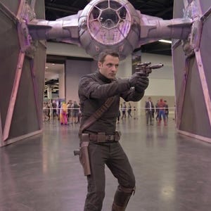 May include: A person in a black costume, holding a gun, stands in front of a large model of a TIE fighter. The scene appears to be at a convention or event, with other people in the background.