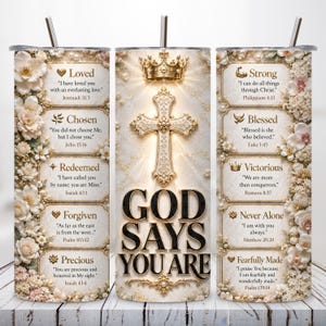 May include: A cylindrical tumbler with a straw, featuring a cream-colored background with white floral accents. The design includes a golden crown, a cross, and the text "GOD SAYS YOU ARE" alongside religious verses like "Loved," "Chosen," and "Strong."