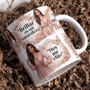 May include: A white ceramic mug featuring illustrations of a woman in a pink dress, surrounded by floral and butterfly motifs. The text reads "Brillar es mi naturaleza" and "Hoy me elijo" in black script. The design includes soft pink roses and bows.
