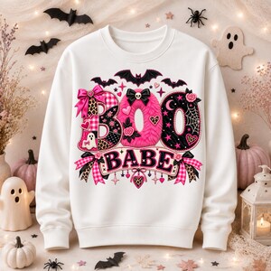 Puede incluir: Una sudadera blanca de cuello redondo con un estampado gráfico que dice BOO BABE. Las letras están decoradas con pelo rosa, telarañas negras, estampado de leopardo, cuadros, lazos, calaveras, murciélagos, corazones y pequeños fantasmas.