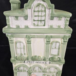 Peut inclure: Un pot à biscuits en céramique en forme de bâtiment à deux étages avec un coloris vert et blanc. La façade présente des fenêtres arquées, une cheminée et le texte POST OFFICE au-dessus de l'entrée. Une petite boîte aux lettres bleue avec le mot MAIL est visible.