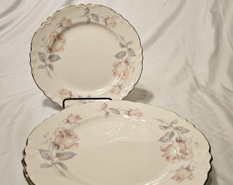May include: A set of three round ceramic plates with a scalloped edge and a gold-tone rim. The plates feature a delicate floral pattern of soft pink and peach roses with muted green and blue-grey leaves arranged along the white surface.