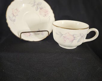 May include: A cream-colored porcelain teacup and matching saucer set featuring a delicate pink rose floral pattern with green leaves. Both pieces are finished with a thin gold trim along the scalloped edges and the rim of the cup.