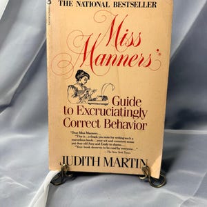 May include: A paperback book titled Miss Manners' Guide to Excruciatingly Correct Behavior by Judith Martin. The cover is light tan with dark red text and a black and white illustration of a person using a typewriter. It rests on a metal stand.