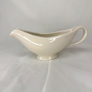 May include: A cream-colored ceramic gravy boat featuring a smooth, glossy finish, a wide pouring spout, a curved handle on the side, and a pedestal base. The vessel has an elongated, oval-shaped body with clean, simple lines and a uniform color.