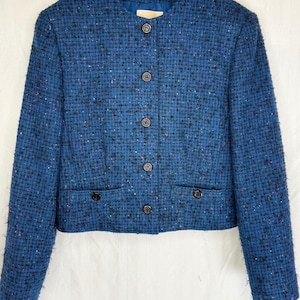 May include: A cropped blue tweed jacket featuring a textured weave with multicolored flecks, a round neckline, long sleeves, and a front button closure with six dark buttons. Two small decorative flap pockets are positioned at the waist.