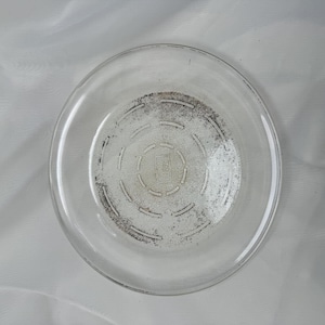 Peut inclure: Une assiette en verre transparent de forme circulaire avec un bord lisse et une base texturée. Le centre de l'assiette présente une série de motifs circulaires en pointillés concentriques et un petit logo rectangulaire en relief au milieu.