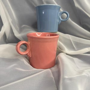 May include: Two ceramic mugs with a smooth, glossy finish. One mug is a soft coral pink colour, and the other is a light periwinkle blue. Both mugs feature a cylindrical body, a wide rim, and a circular handle attached to the side of the vessel.