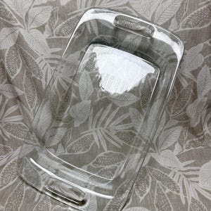 May include: A clear, rectangular baking dish with handles. The dish is made of transparent glass and has a smooth surface. The handles are integrated into the design, providing a secure grip. The dish is designed for baking and serving.