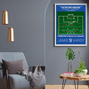 Op de afbeelding: Een ingelijste poster met een blauwe rand toont een voetbalveld diagram. Tekst bovenaan luidt "THE RECORD BREAKER" en geeft details over de wedstrijd Leicester City tegen Manchester United. Daaronder staat "VARDY!!!It's Eleven, It's Heaven" en "JAMIE 9 VARDY".