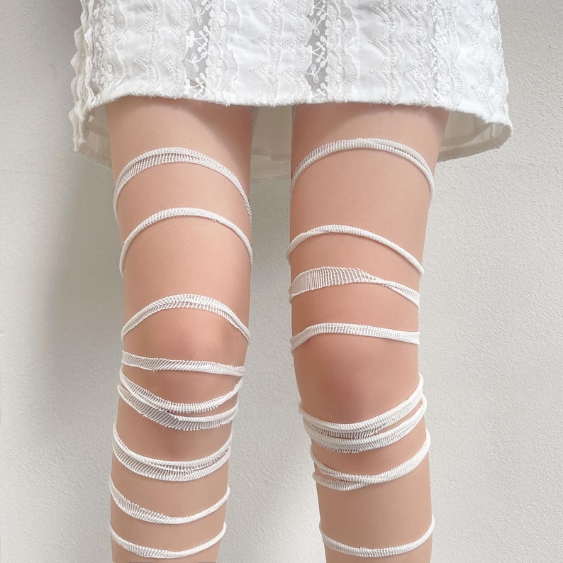 May include: A white lace skirt paired with white leg wraps. The leg wraps consist of multiple thin, horizontal bands of white fabric that encircle the legs from the mid-thigh down to the lower calf, creating a layered, striped appearance.