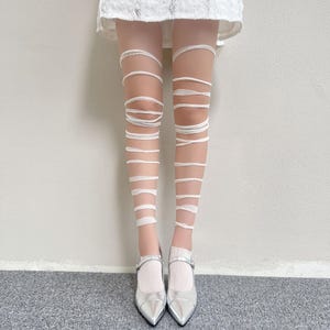 May include: Sheer white tights featuring a horizontal wrap-around bandage design that extends from the upper thigh to the ankle, paired with metallic silver pointed-toe Mary Jane flats and a white lace hem visible at the top of the frame.
