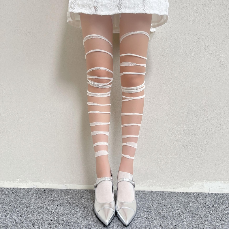 May include: Sheer white tights featuring a horizontal wrap-around bandage design that extends from the upper thigh to the ankle, paired with metallic silver pointed-toe Mary Jane flats and a white lace hem visible at the top of the frame.