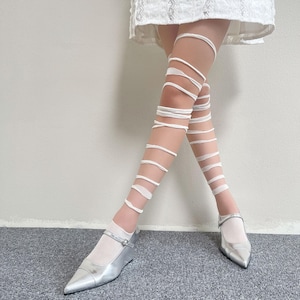 May include: White leg wraps featuring a spiral design that extends from the ankle to the upper thigh, paired with metallic silver pointed-toe shoes with a single strap and a white skirt hem visible at the top of the frame.
