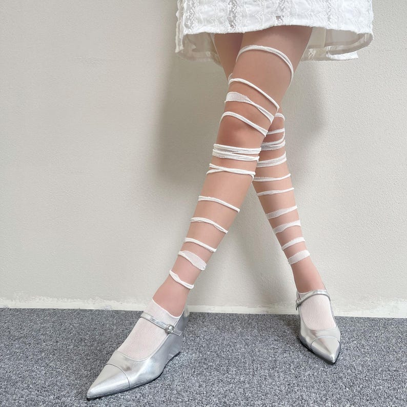 May include: White leg wraps featuring a spiral design that extends from the ankle to the upper thigh, paired with metallic silver pointed-toe shoes with a single strap and a white skirt hem visible at the top of the frame.
