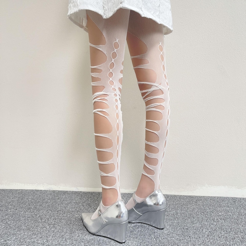 May include: White tights featuring a distressed, laddered, and cut-out pattern that runs vertically along the legs. The hosiery is paired with metallic silver wedge shoes and a white skirt, creating a layered look with visible skin through the gaps.