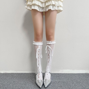 May include: White lace knee-high socks with a ruffled top and ribbon ties. The socks are worn with silver pointed-toe heels with a strap. The image shows a fashion outfit.