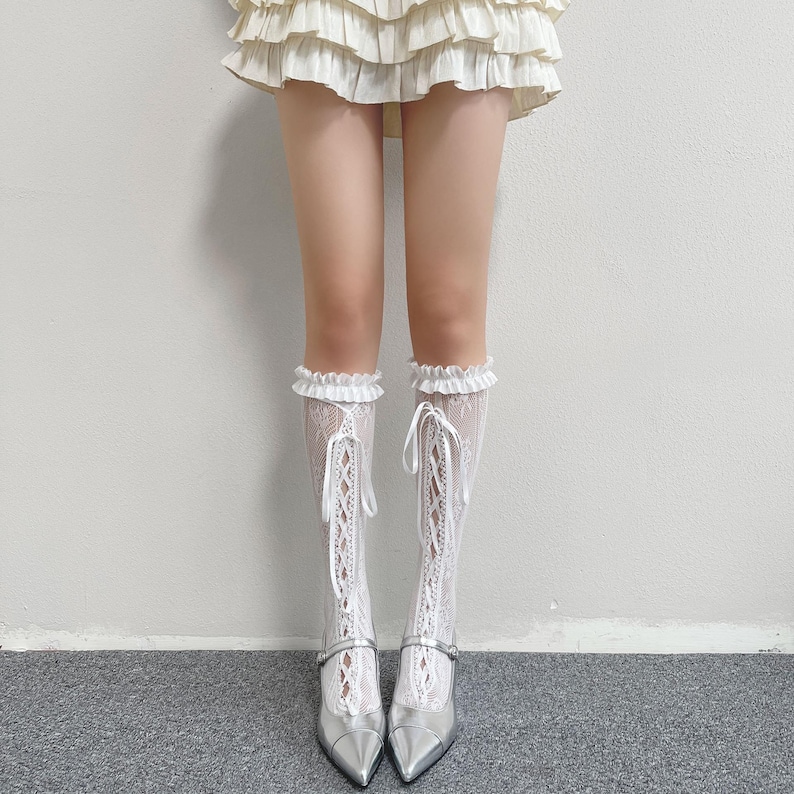 May include: White lace knee-high socks with a ruffled top and ribbon ties. The socks are worn with silver pointed-toe heels with a strap. The image shows a fashion outfit.
