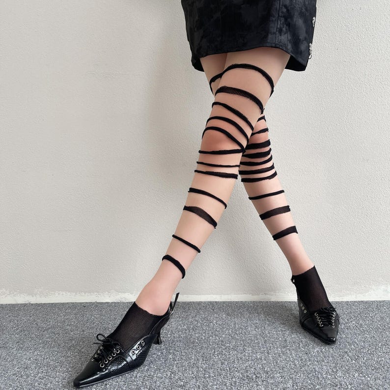 May include: Sheer nude tights featuring a series of horizontal black fabric bands that wrap around the legs from the upper thigh down to the ankle. The tights are paired with black pointed-toe heels that have lace-up detailing and a short heel.