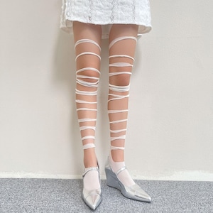 May include: White leg wraps featuring a series of horizontal bands that extend from the ankle to the upper thigh, paired with metallic silver wedge shoes with a pointed toe and a single strap across the top of the foot, worn under a white skirt.