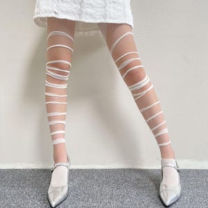 May include: Sheer nude tights featuring a white wrap-around bandage design that spirals from the upper thigh down to the ankle, paired with metallic silver pointed-toe Mary Jane shoes and a white skirt hem visible at the top of the frame.