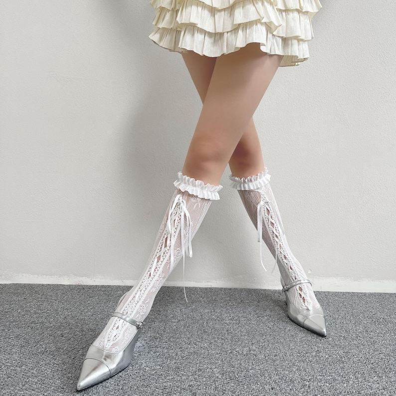May include: White lace knee-high socks with a frilled top and ribbon ties. The socks are worn with silver pointed-toe heels. The overall aesthetic is delicate and feminine, suitable for special occasions or adding a touch of elegance to an outfit.