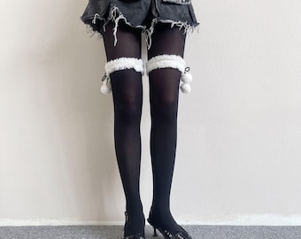 May include: A dark gray distressed denim mini skirt with frayed edges, worn over opaque black tights. The tights feature white fuzzy leg warmers with dangling pom-poms at the thighs. Black pointed-toe slingback heels with lace-up details.